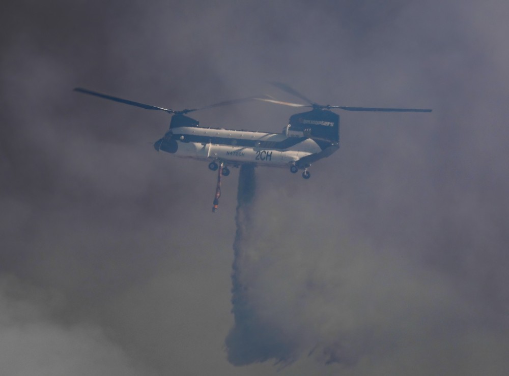 PJ Helicopters Helitanker 2CH, operating in the San Diego County (CA) area during a rapidly spreading wildfire, dumps swamp water on the flames below during the Garden Fire in Fallbrook, CA.