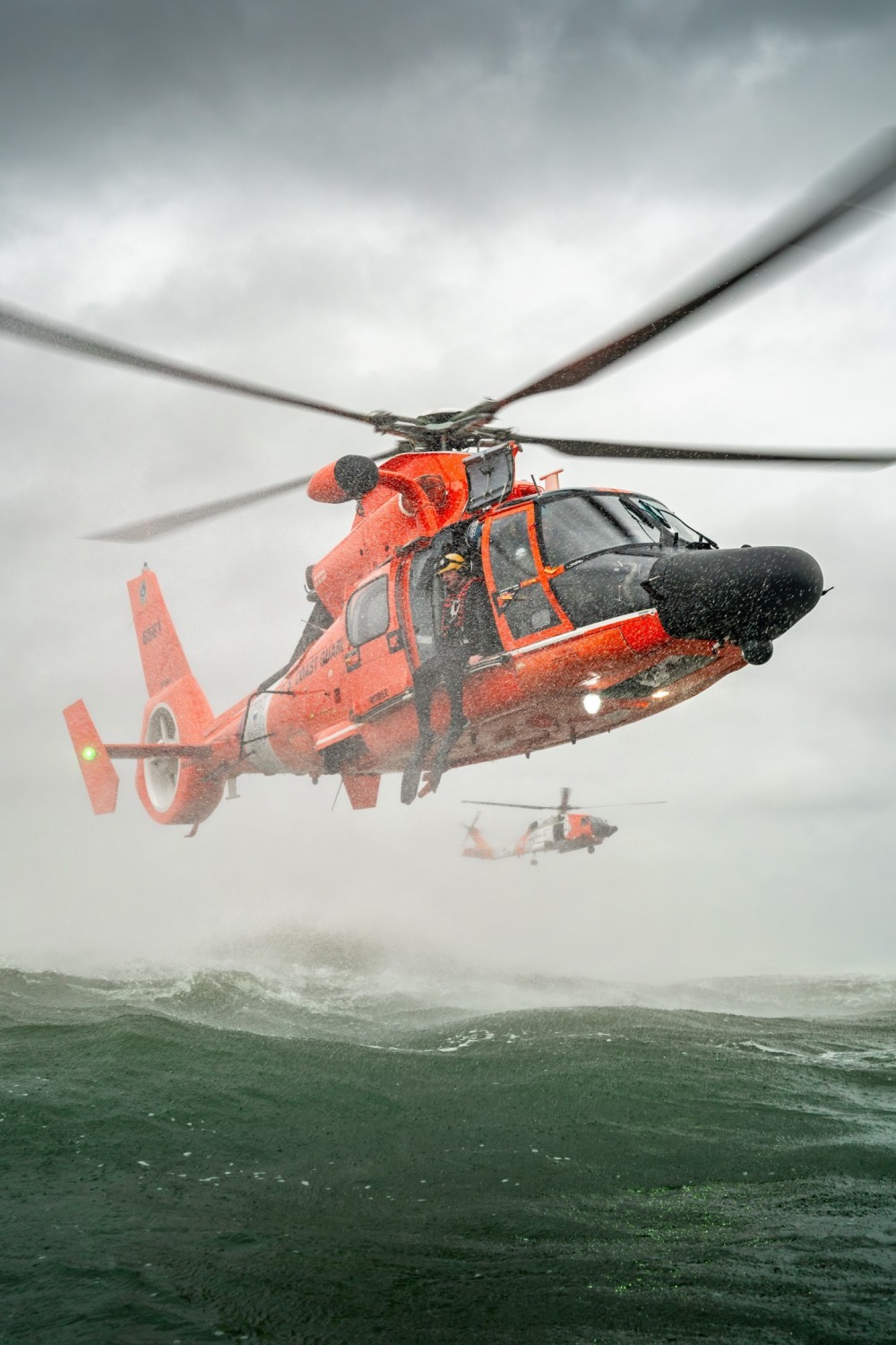 A Coast Guard Rescue Swimmer prepares for a Free Fall Deployment from a MH-65E during a training exercise while a MH-60T is looking from afar.